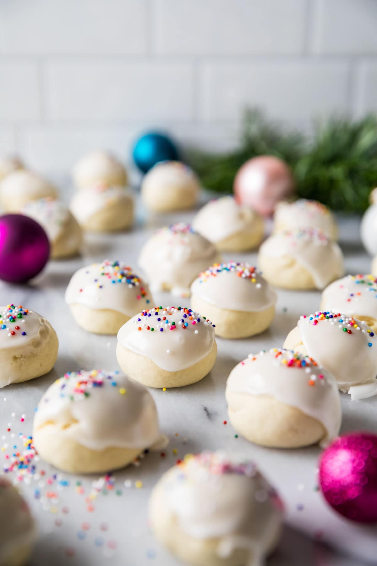 Round cookies topped with a white icing and rainbow nonpareil sprinkles with colorful Christmas ornaments mixed in.
