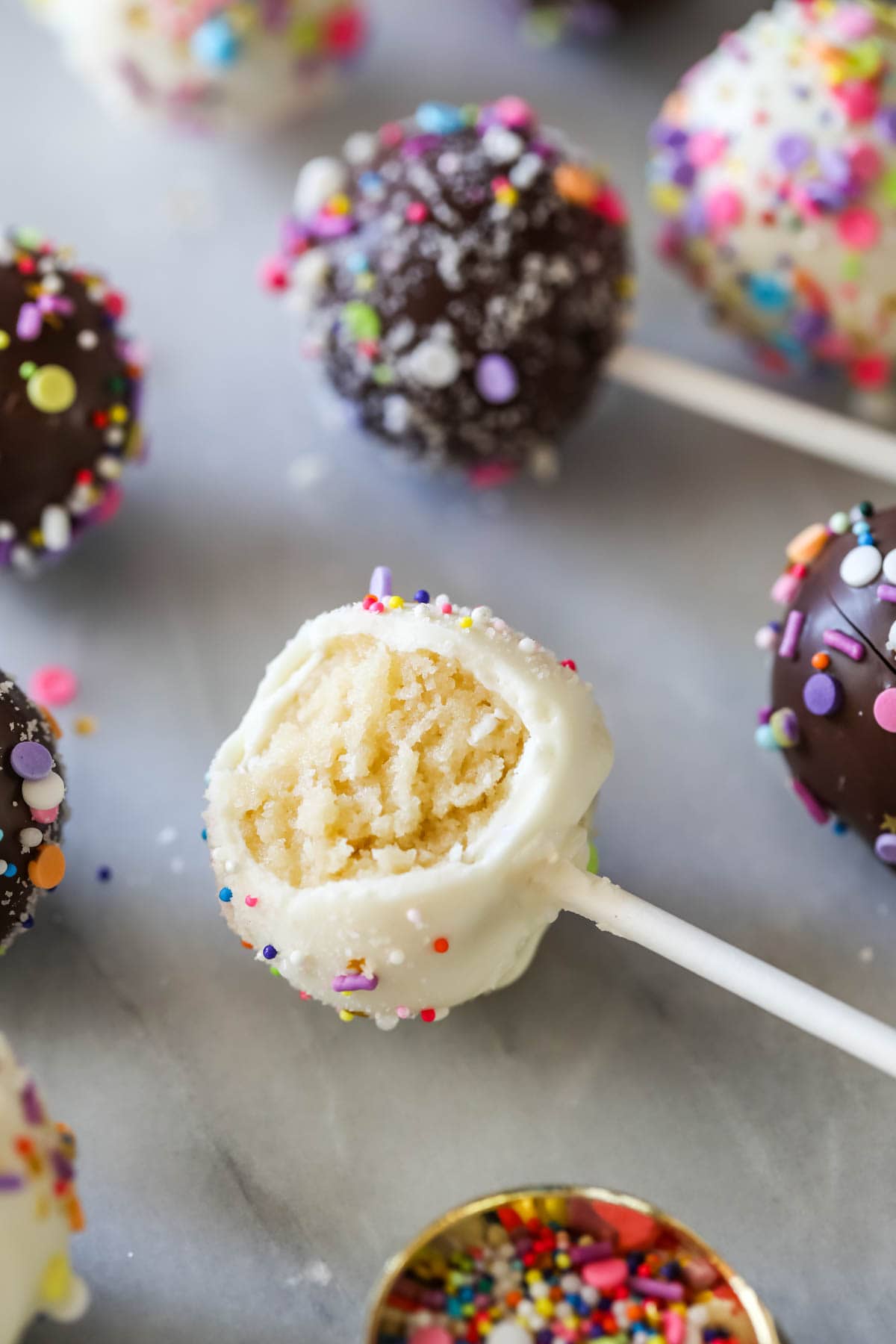 Candy coated cake balls on sticks with one bitten to show a yellow cake interior.
