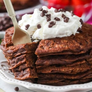 Fork taking a bite out of a stack of chocolate pancakes topped with whipped cream and chocolate chips.