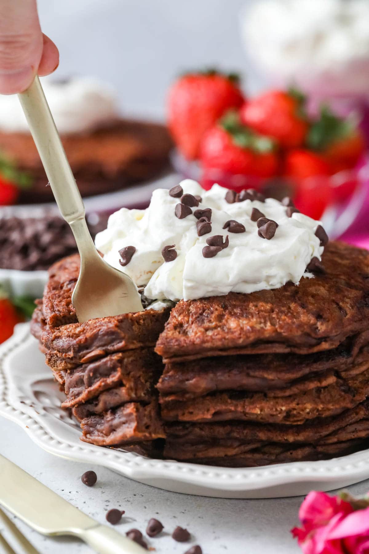 Fork taking a bite out of a stack of chocolate pancakes topped with whipped cream and chocolate chips.