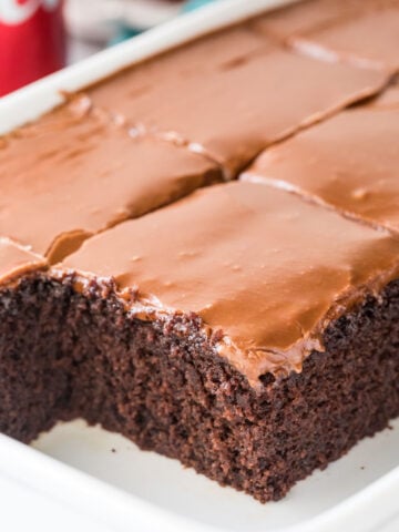 Thick squares of coca cola cake with shiny brown frosting in white cake pan