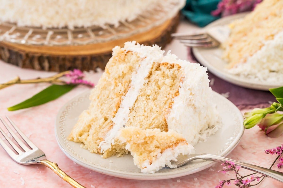Slice of coconut cake on white plate with forkful missing. Pink background with flowers.
