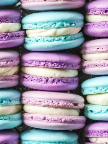 French macaron recipe showing side view of bright blue, light purple, and dark purple macarons sandwiches with white filling, all arranged in neat rows