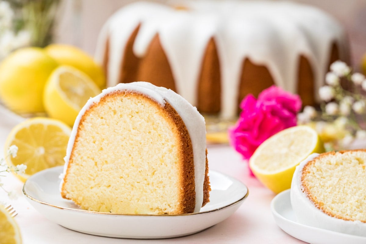 Lemon pound cake slice on a white plate with lemons and whole cake in the background.