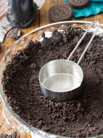 Measuring cup pressing oreo cookie crumbs into bottom of pie plate