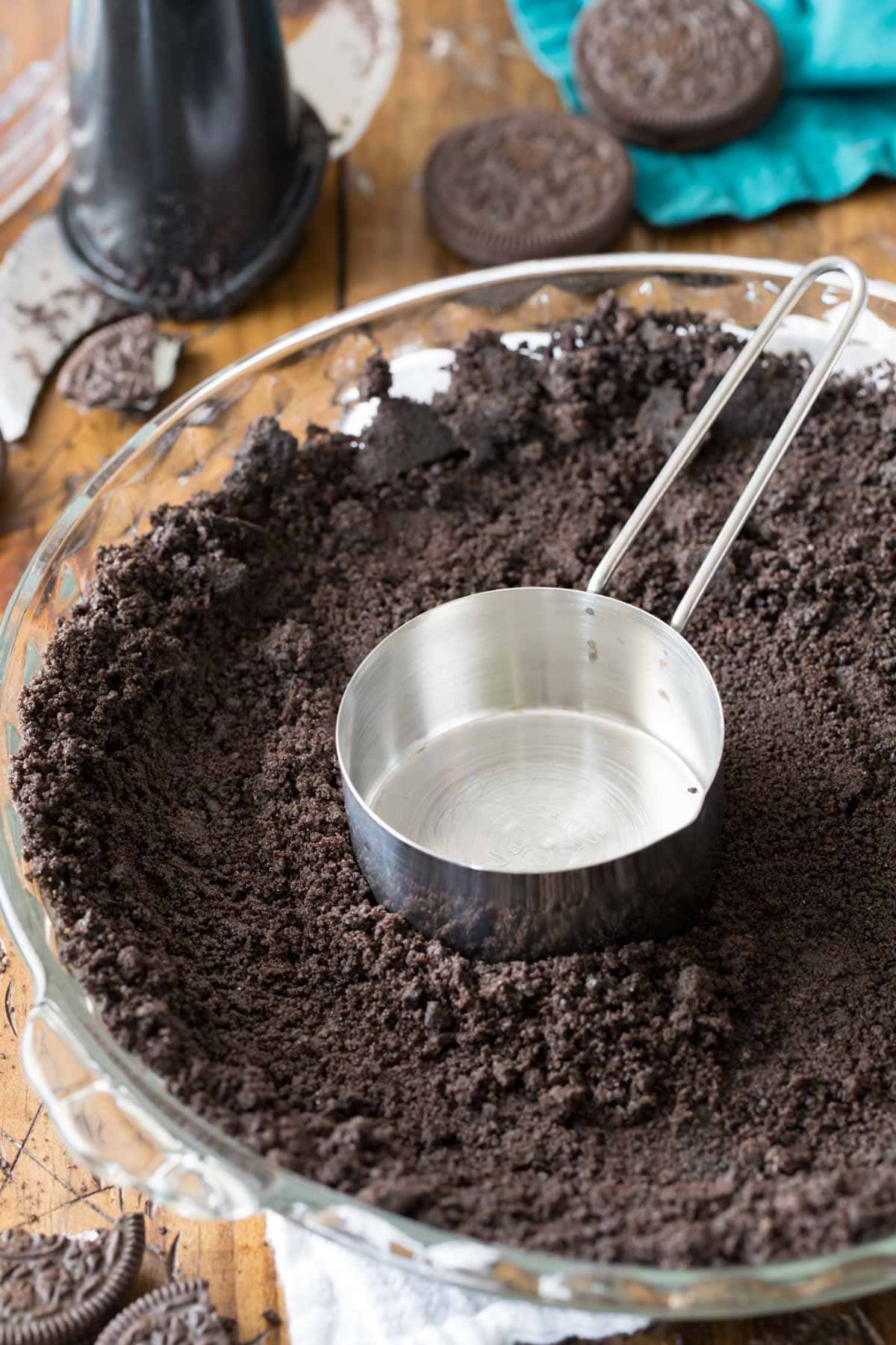 Metal measuring cup tamping down Oreo crumbs into a pie plate to form a crust.