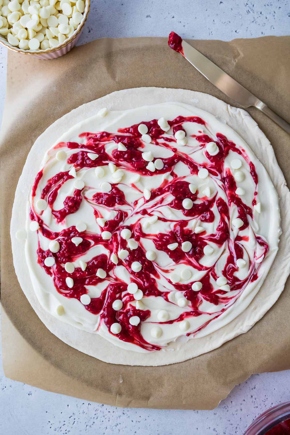 Overhead view of pizza dough topped with a swirled raspberry cream cheese sauce and white chocolate chips.