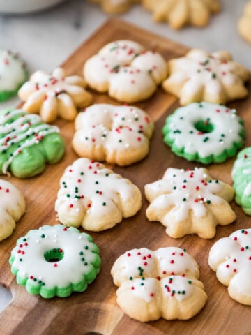 Colorful and festive spritz cookies shaped like wreaths, trees, and snowflakes on a wood cutting board.