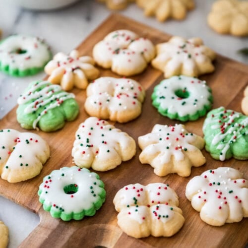 Colorful and festive spritz cookies shaped like wreaths, trees, and snowflakes on a wood cutting board.