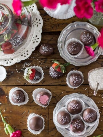 Overhead of strawberry buttercream candies arranged on a silver plate and wood surface, surrounded by chocolate covered strawberries