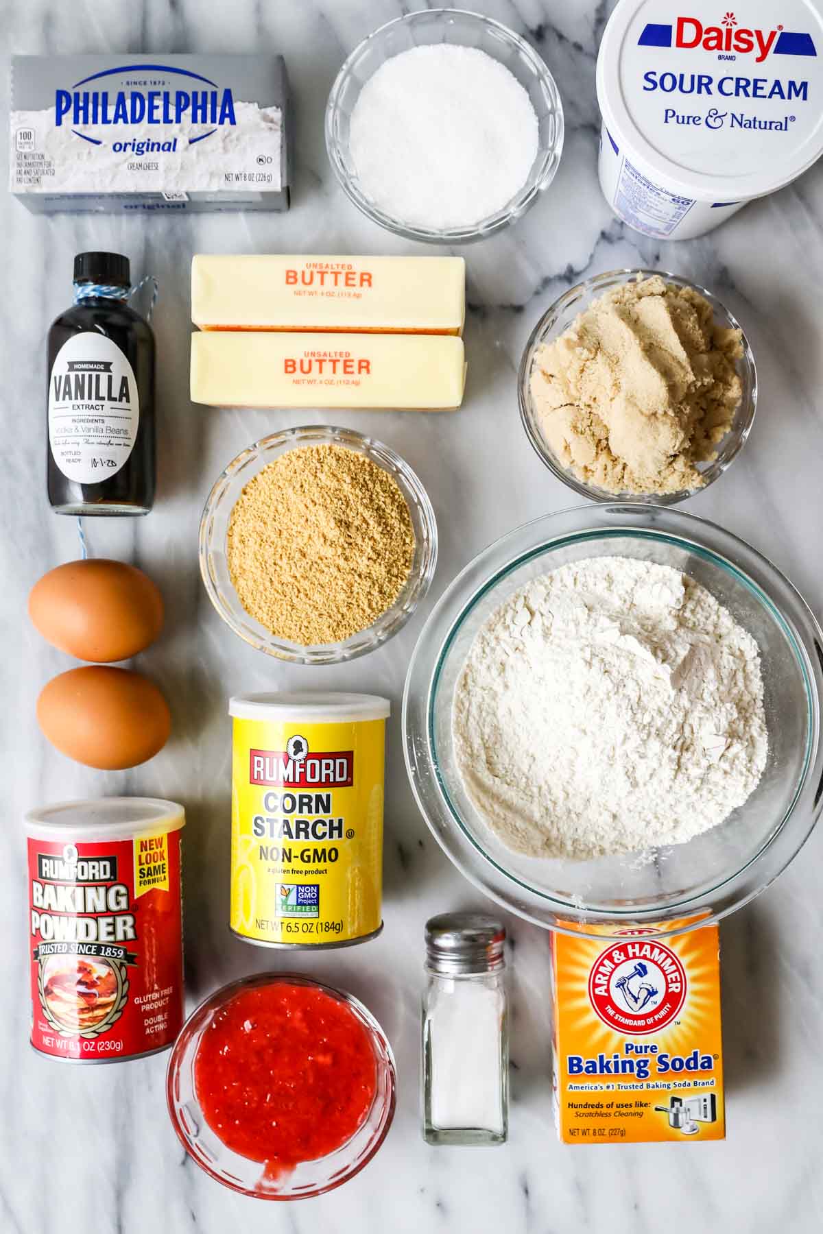 Overhead view of ingredients including graham cracker crumbs, cream cheese, strawberry sauce, and more.