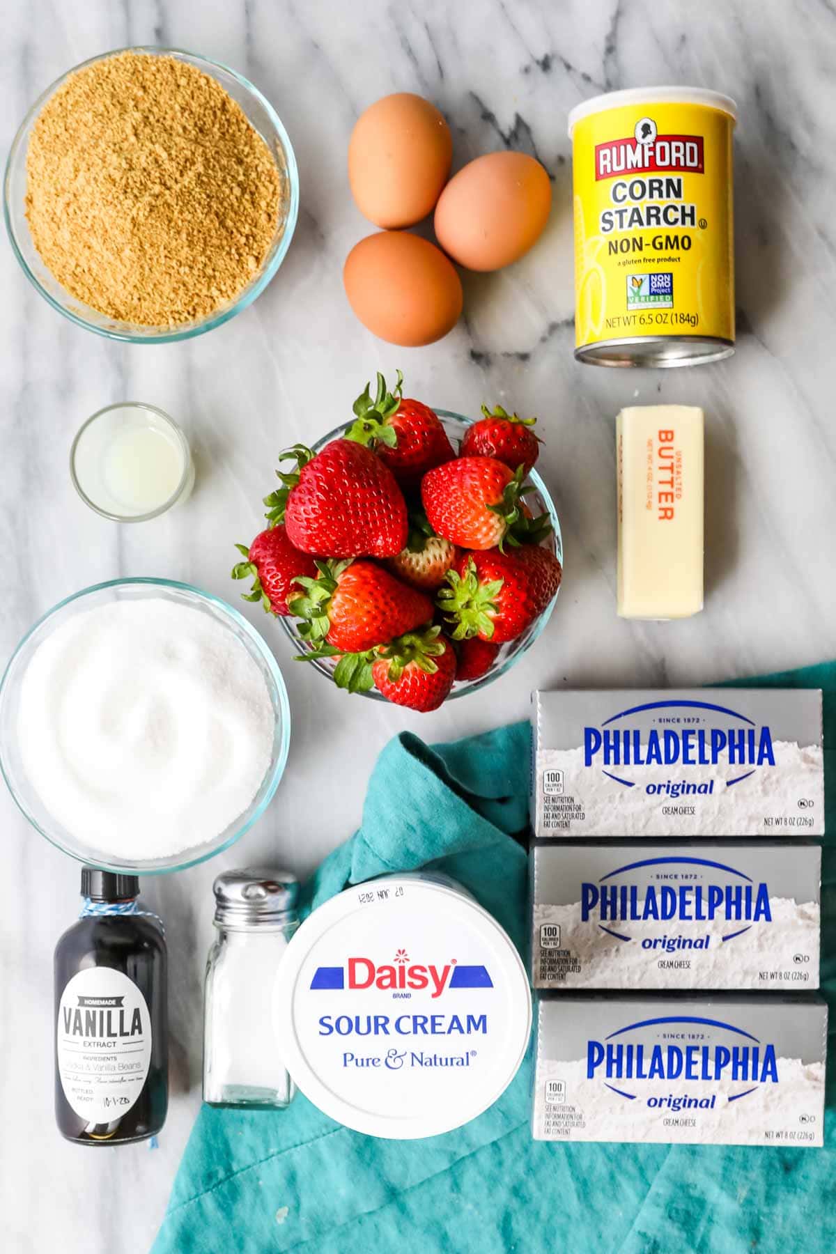 Overhead view of ingredients including graham cracker crumbs, strawberries, cream cheese, sour cream, and more.